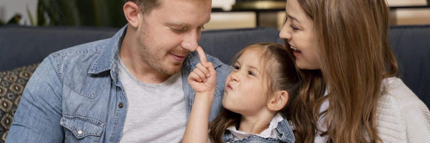Uomo e donna seduti su un divano con una bambina tra loro. La bambina tocca il naso dell'uomo con il dito mentre la donna la osserva sorridendo. Scena di interazione familiare in ambiente domestico.