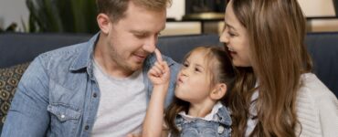 Uomo e donna seduti su un divano con una bambina tra loro. La bambina tocca il naso dell'uomo con il dito mentre la donna la osserva sorridendo. Scena di interazione familiare in ambiente domestico.