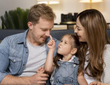 Uomo e donna seduti su un divano con una bambina tra loro. La bambina tocca il naso dell'uomo con il dito mentre la donna la osserva sorridendo. Scena di interazione familiare in ambiente domestico.