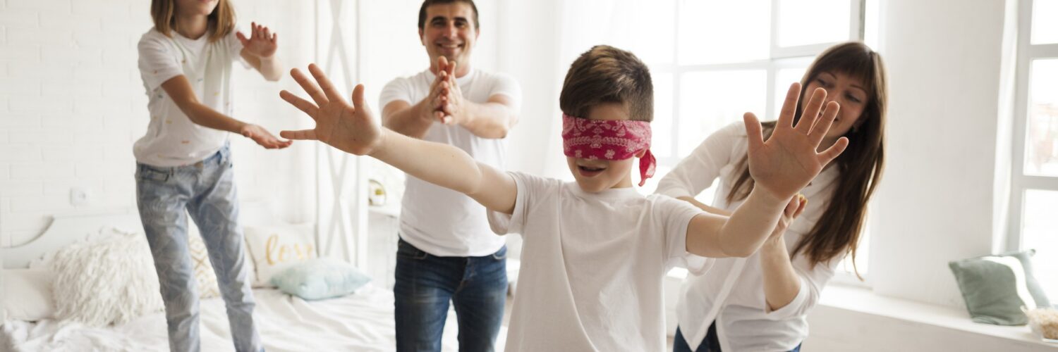 Una famiglia di quattro persone gioca allegramente a mosca cieca in una camera da letto luminosa. Al centro, un ragazzo bendato protende le braccia, mentre i genitori e la sorella gli si muovono intorno sorridendo per non farsi acchiappare.