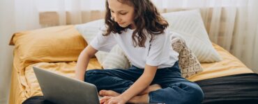 bambina che studia a casa seduta sul letto con davanti un computer e un quaderno con poggiata su una penna