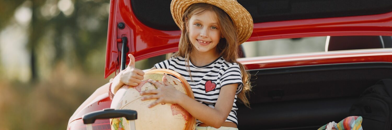 Una bambina seduta nel bagagliaio aperto di un’auto rossa, sorridente, con indosso un cappello di paglia, una maglietta a righe bianche e nere e pantaloncini chiari. Tiene in braccio un grande mappamondo e fa il gesto del pollice in su. Accanto a lei ci sono una valigia con manico estensibile e una borsa colorata a righe. La scena si svolge all’aperto, in un contesto estivo.