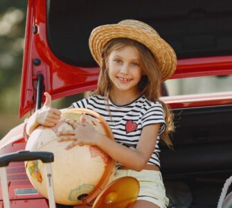 Una bambina seduta nel bagagliaio aperto di un’auto rossa, sorridente, con indosso un cappello di paglia, una maglietta a righe bianche e nere e pantaloncini chiari. Tiene in braccio un grande mappamondo e fa il gesto del pollice in su. Accanto a lei ci sono una valigia con manico estensibile e una borsa colorata a righe. La scena si svolge all’aperto, in un contesto estivo.