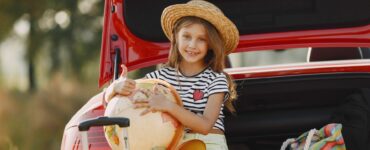 Una bambina seduta nel bagagliaio aperto di un’auto rossa, sorridente, con indosso un cappello di paglia, una maglietta a righe bianche e nere e pantaloncini chiari. Tiene in braccio un grande mappamondo e fa il gesto del pollice in su. Accanto a lei ci sono una valigia con manico estensibile e una borsa colorata a righe. La scena si svolge all’aperto, in un contesto estivo.
