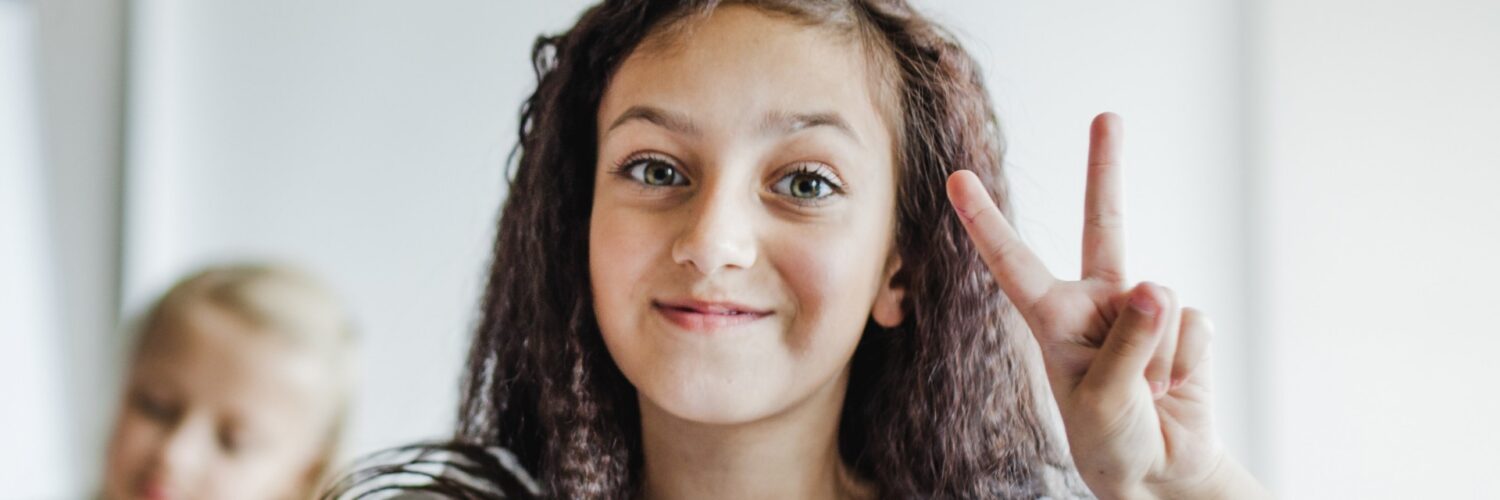 Una bambina con i capelli ricci e scuri sorride seduta a un banco di scuola, all'interno di un'aula luminosa, facendo il segno di vittoria con la mano sinistra.
