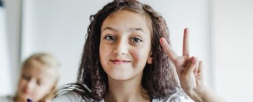 Una bambina con i capelli ricci e scuri sorride seduta a un banco di scuola, all'interno di un'aula luminosa, facendo il segno di vittoria con la mano sinistra.