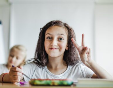 Una bambina con i capelli ricci e scuri sorride seduta a un banco di scuola, all'interno di un'aula luminosa, facendo il segno di vittoria con la mano sinistra.